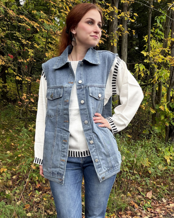 A woman wearing a light blue denim vest over a white long-sleeve top with black-and-white striped cuffs and hem, paired with blue jeans, standing outdoors in a wooded area with autumn foliage.