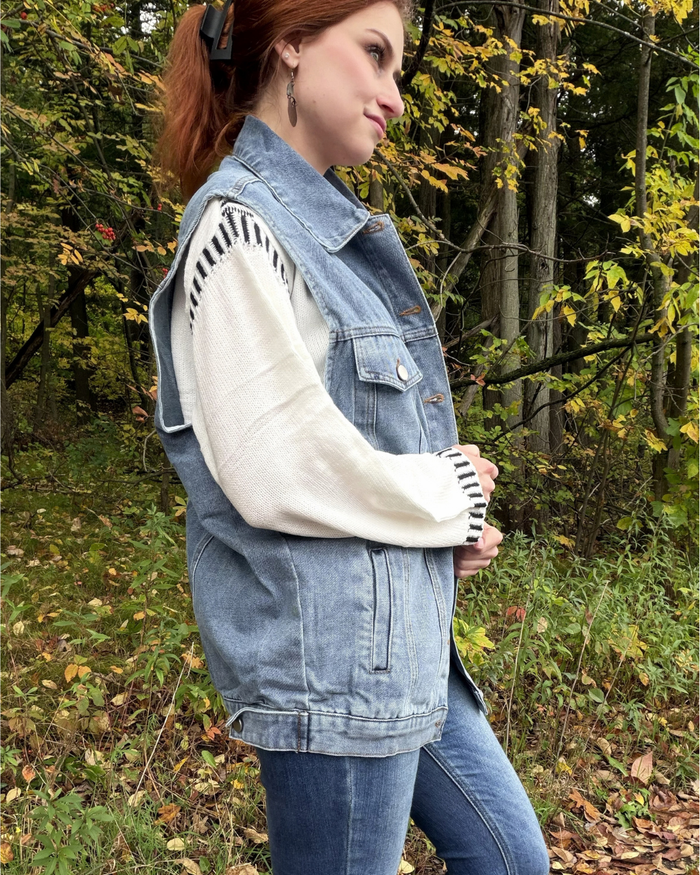 A woman wearing a light blue denim vest over a cream knit sweater with black-and-white striped sleeve details, paired with blue jeans, standing outdoors in a wooded area with autumn foliage.