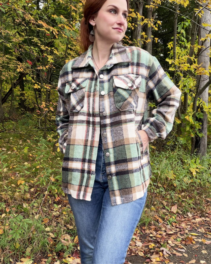 A woman wearing a green, beige, and brown plaid flannel button-up shirt jacket over a blue denim shirt and jeans, standing outdoors in a wooded area with autumn foliage.