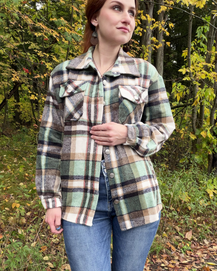 A woman wearing a vintage-style green, beige, and brown plaid flannel shirt jacket over a black top and blue jeans, standing outdoors in a wooded area with autumn foliage.