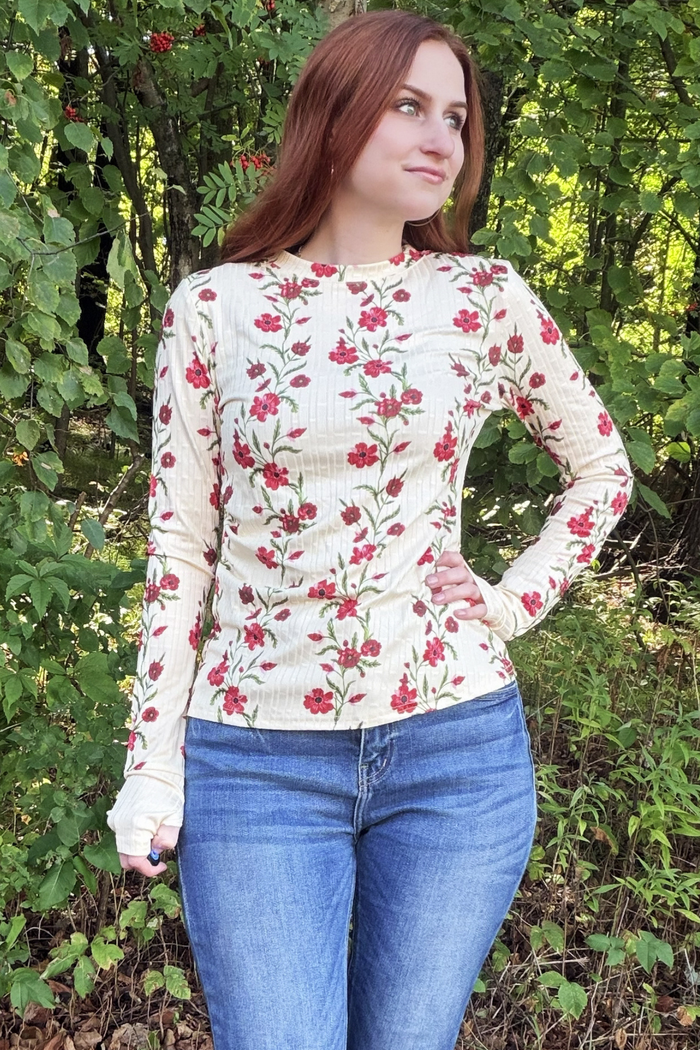 A woman wearing a cream-colored long-sleeve top with a red and green floral pattern, paired with blue jeans, standing outdoors in front of green foliage.