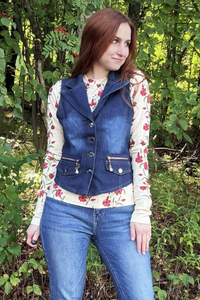 A woman wearing a dark blue denim vest over a cream floral long-sleeve shirt and blue jeans, standing outdoors in a wooded area with green foliage.
