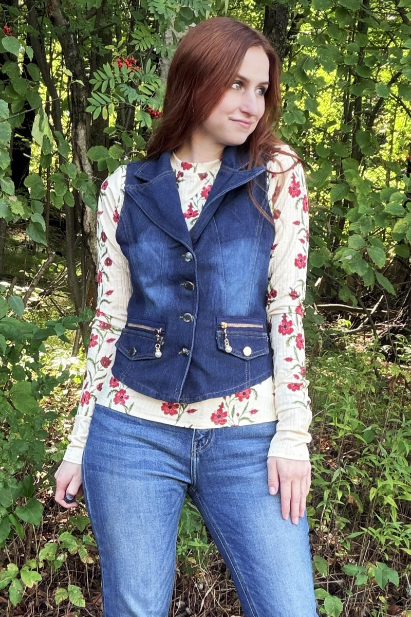 A woman wearing a dark blue denim vest over a cream floral long-sleeve shirt and blue jeans, standing outdoors in a wooded area with green foliage.