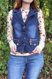 A person outdoors wearing a dark blue denim vest over a cream floral long-sleeve top and blue jeans, standing in front of green foliage.