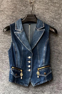 A blue denim vest with a blazer-style collar, decorative gold-tone buttons, and zippered flap pockets with dangling charms, hanging on a black hanger against a gray textured background.
