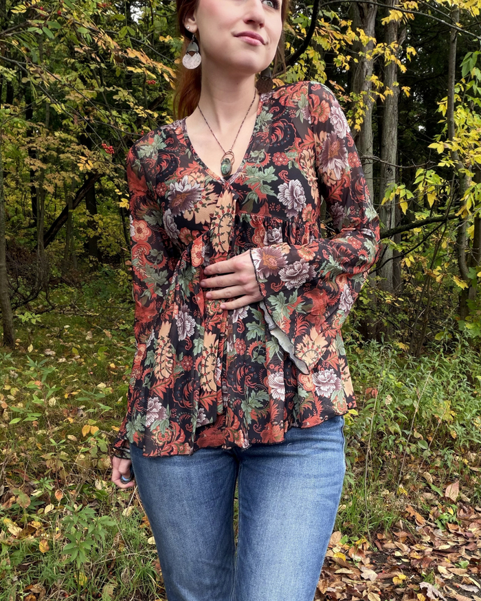 A woman wearing a long-sleeve floral blouse with blue jeans, standing outdoors in a wooded area with autumn foliage.