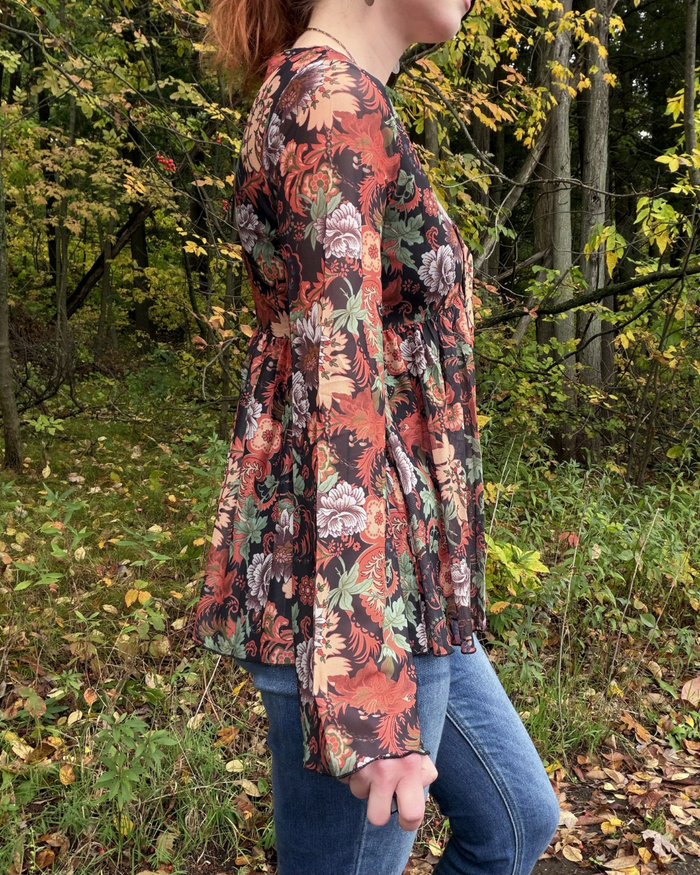 A woman wearing a long-sleeve peplum top with a dense floral and botanical print in rust orange, black, green, and cream, paired with blue jeans, standing in a wooded outdoor setting.