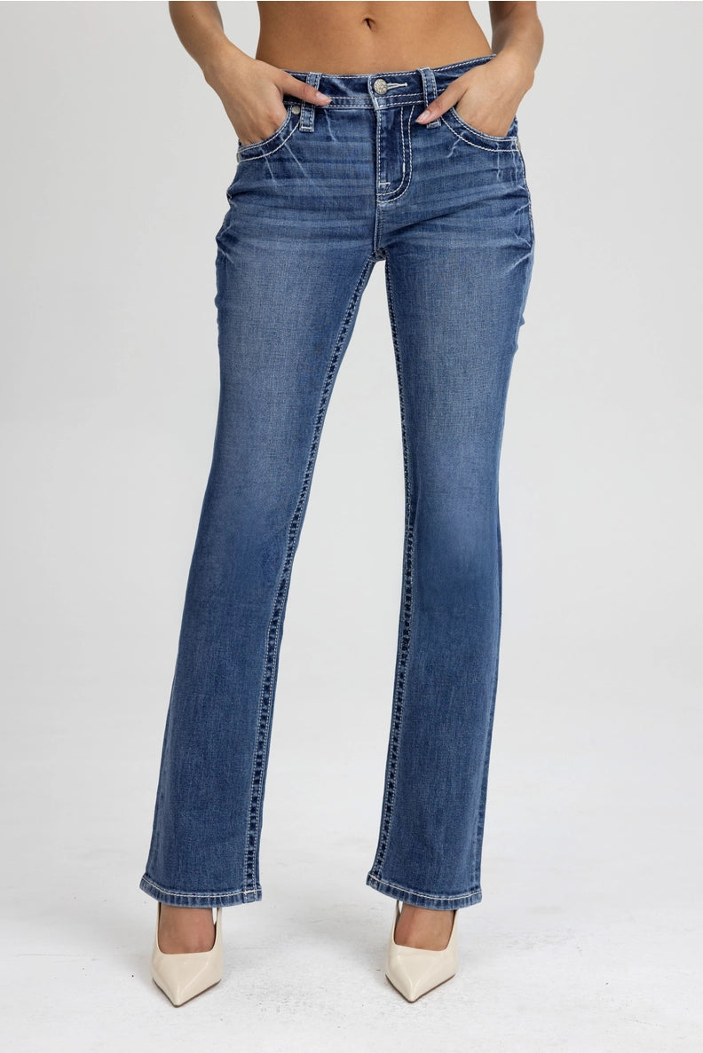 A model wearing a pair of medium-wash blue flare jeans with front pleats and light beige pointed-toe heels, shown from the waist down against a light gray studio background.