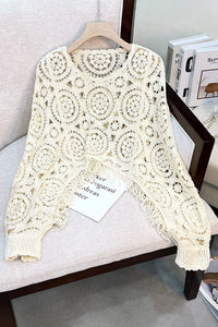 Cream crochet knit sweater with intricate circular lace patterns, displayed on a wooden hanger on a light beige upholstered chair.