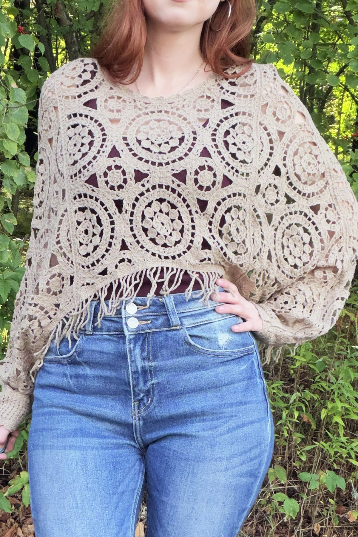 A woman wearing a beige crochet poncho sweater with fringe and blue jeans, standing outdoors in front of green foliage.