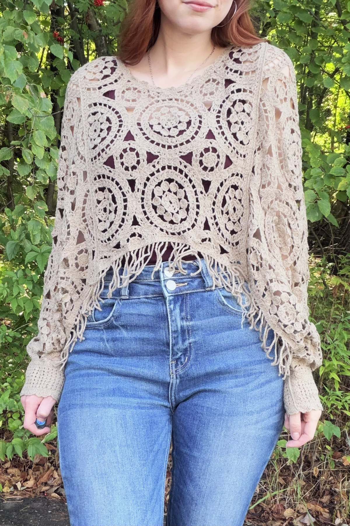 A woman wearing a beige crochet poncho sweater with fringe details, paired with blue jeans, standing outdoors in front of a green leafy background.