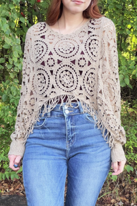A woman wearing a beige crochet poncho sweater with fringe details, paired with blue jeans, standing outdoors in front of a green leafy background.