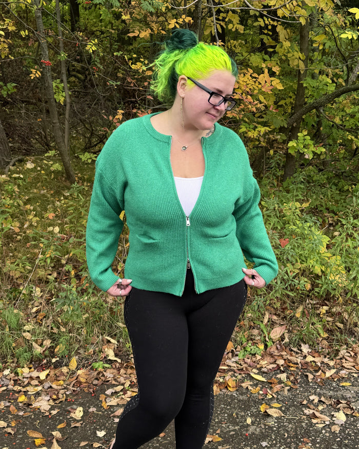 A woman with bright neon green hair and glasses stands outdoors in a wooded area with fallen leaves, wearing a green zip-up cardigan over a white top and black leggings.