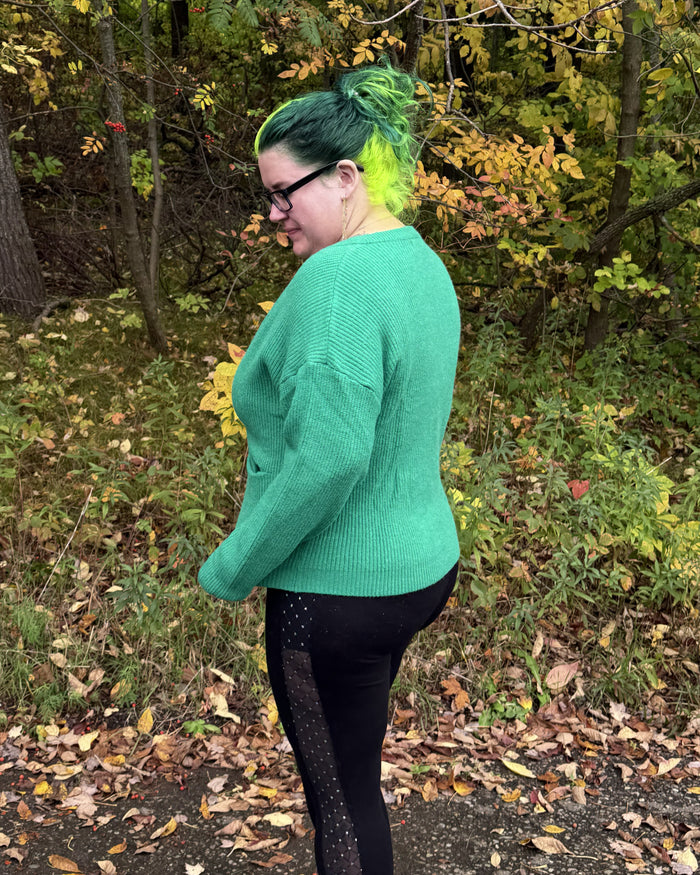 A person standing outdoors in a wooded area with fallen leaves, wearing a bright green ribbed knit sweater and black leggings with dotted side panels.