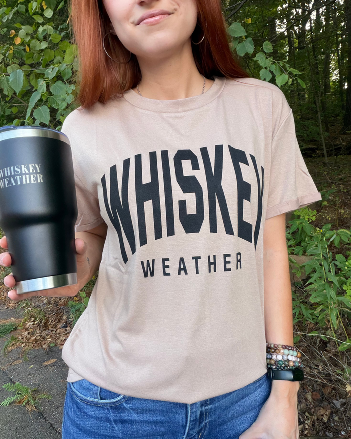 A woman outdoors wearing a beige graphic t-shirt with 'WHISKEY WEATHER' printed in bold black letters, holding a black insulated tumbler with the same text.